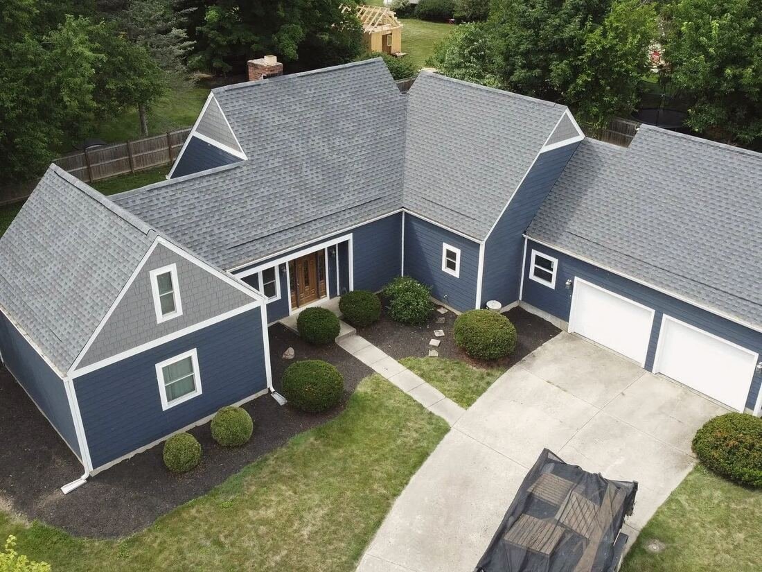 Drone view of a blue-sided home with a new grey shingle roof and a covered trailer in the driveway