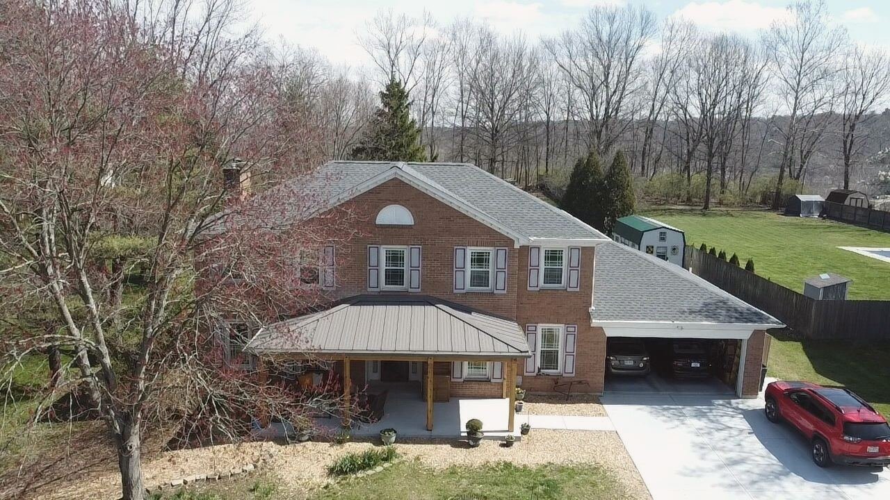 Drone view of a two-story brick home with a grey shingle roof and a standing seam metal porch roof