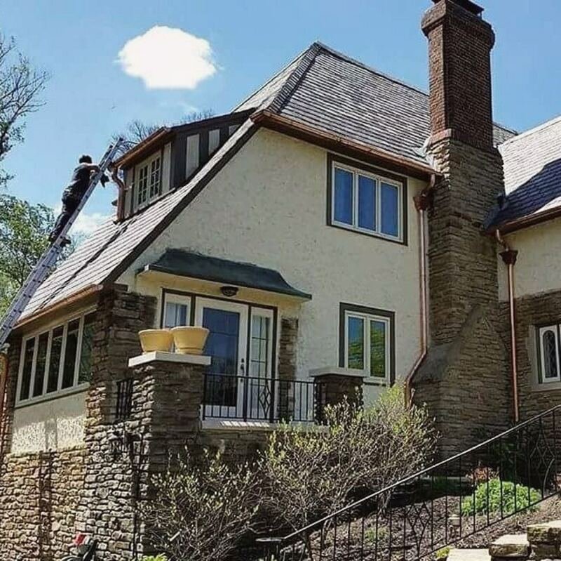 Stucco and stone home with a steep new dark roof, worker on the roof at left