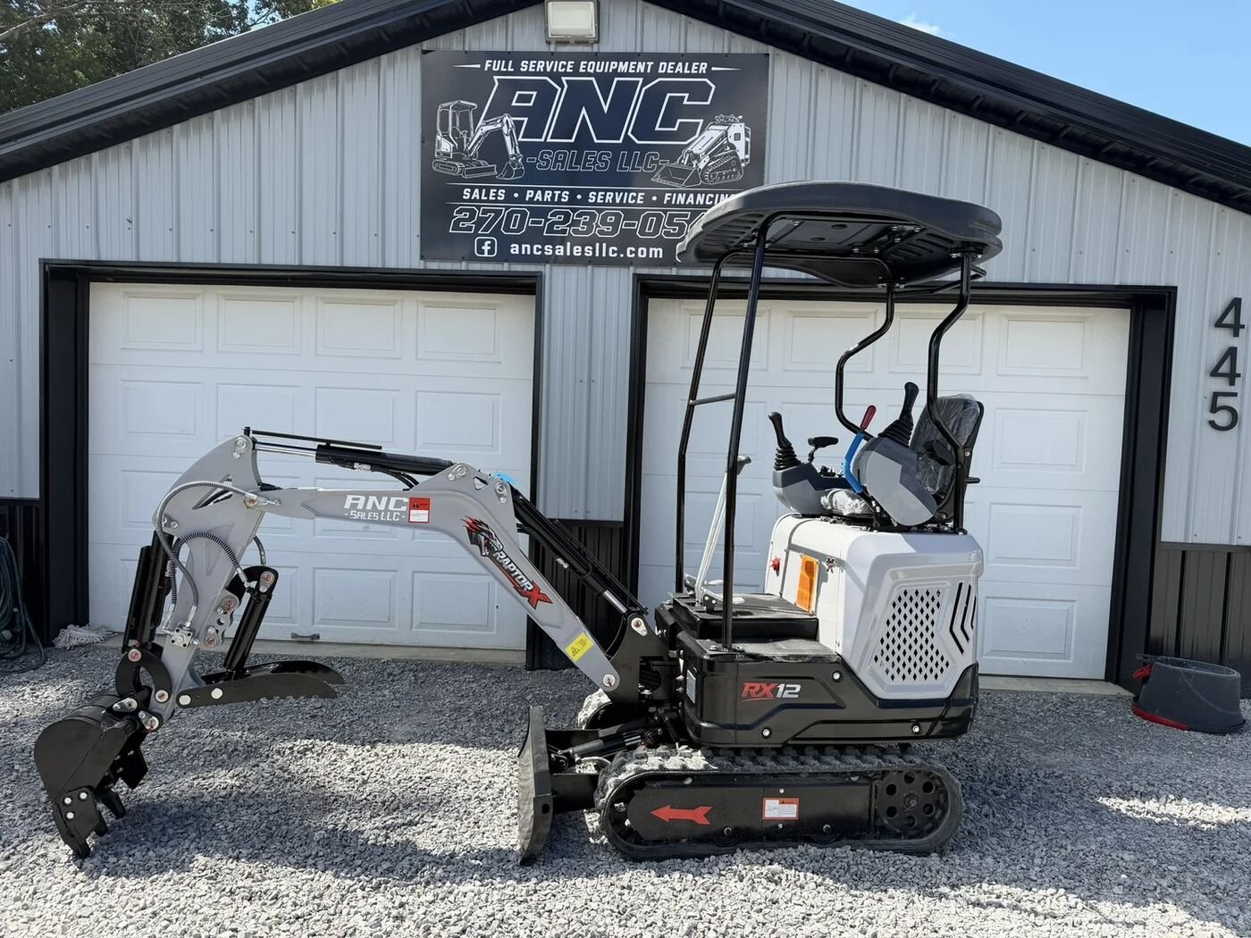 Gray and white mini excavator with its bucket curled, parked on gravel in front of a metal shop building with an ANC Sales sign