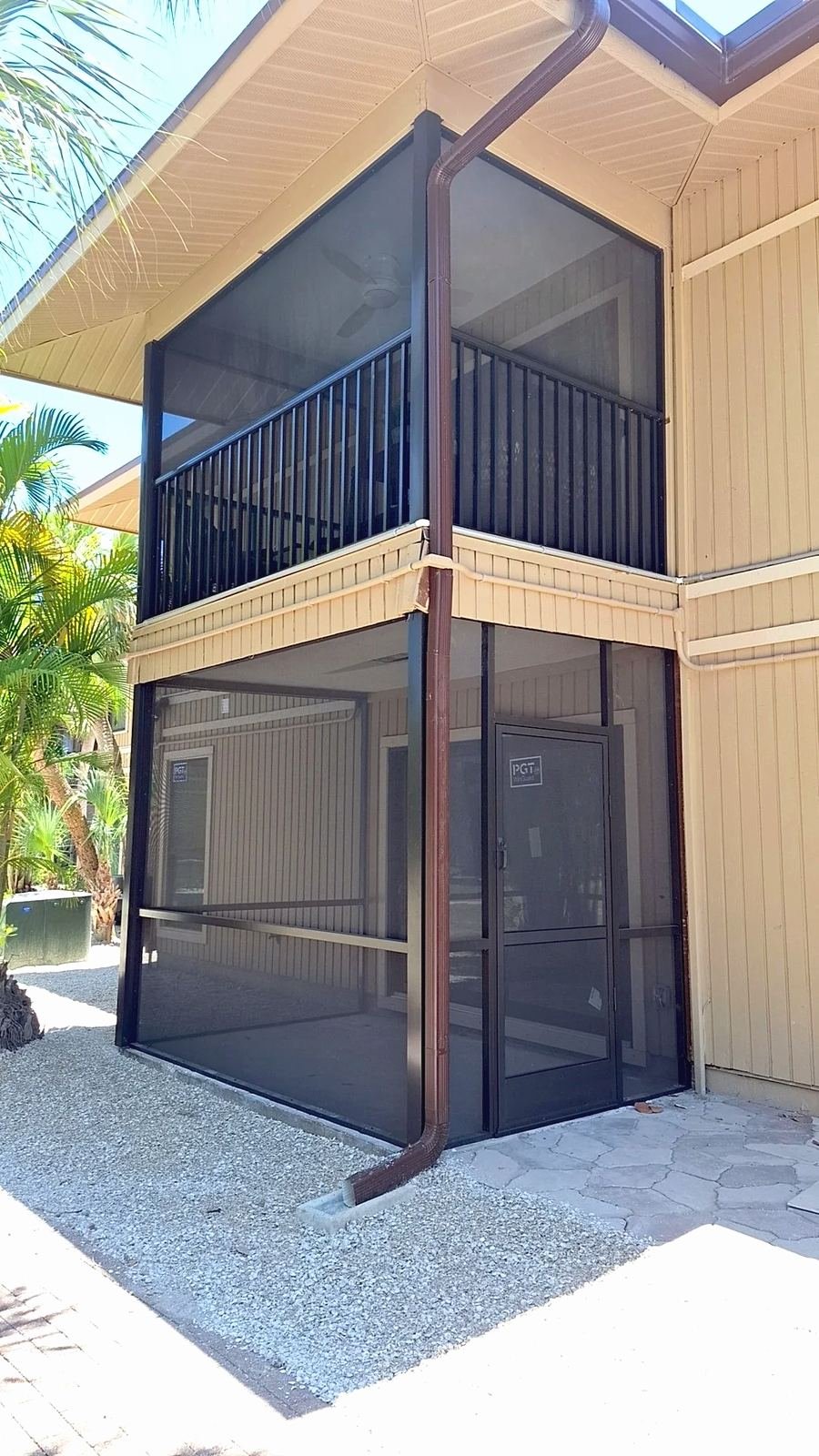 Two-story bronze aluminum screen enclosure on a tan stilt house, black balcony railing above, gravel and pavers below