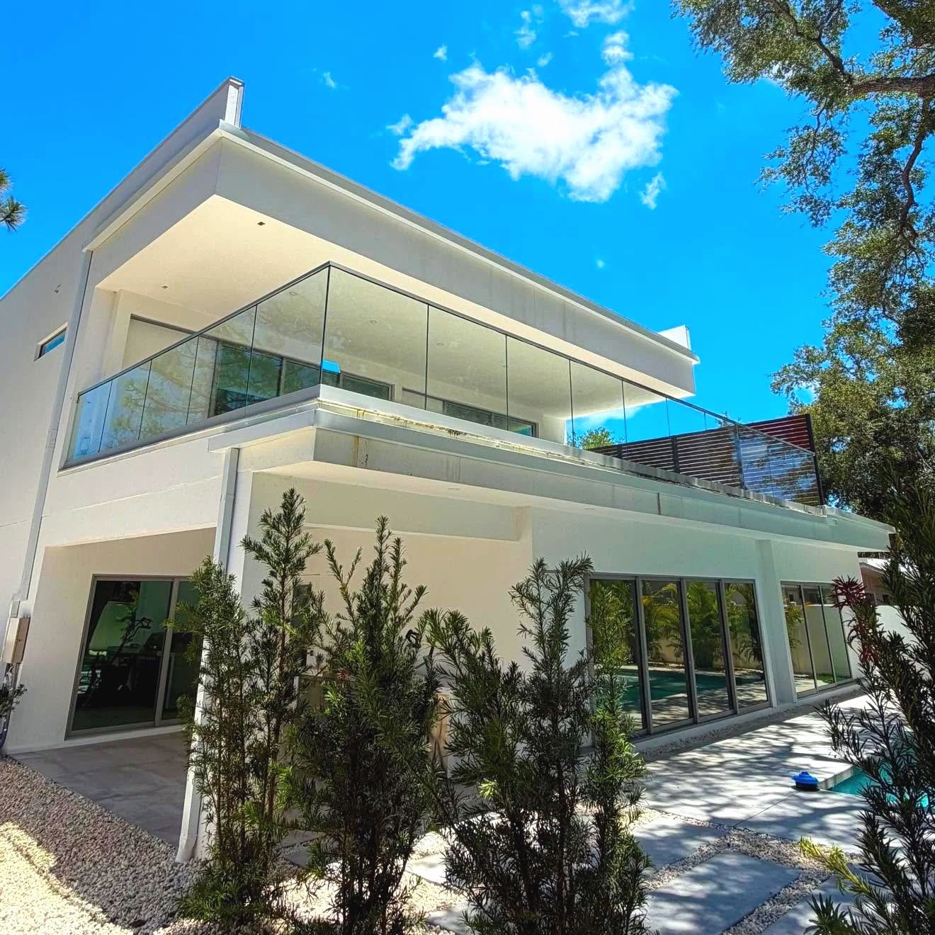Two-story white modern house with a frameless glass balcony rail and wood-look slat cladding accent, shrubs and a pool at the side