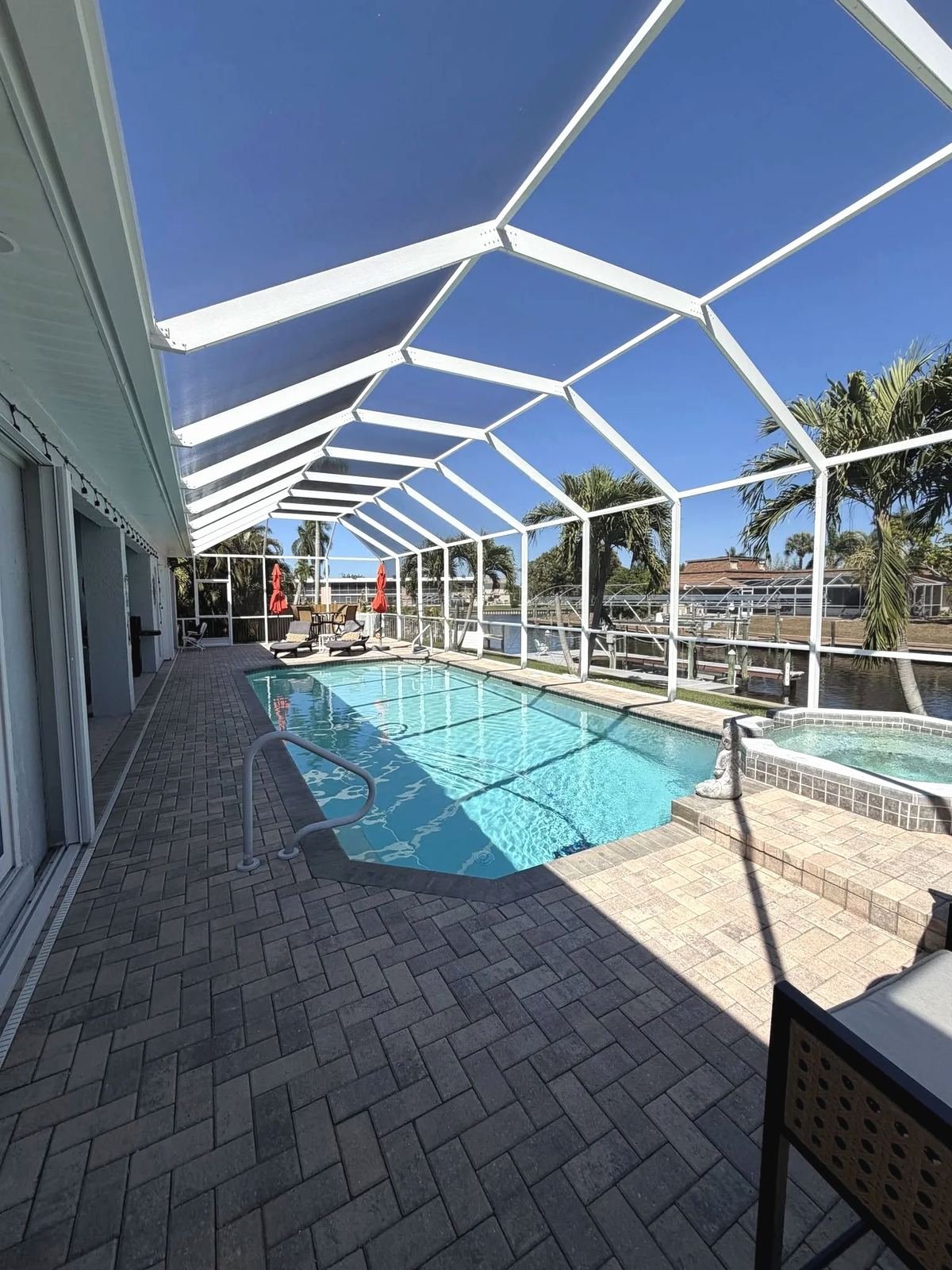 Inside a restored white pool cage looking down a lap pool and spa toward the water, paver deck in the foreground