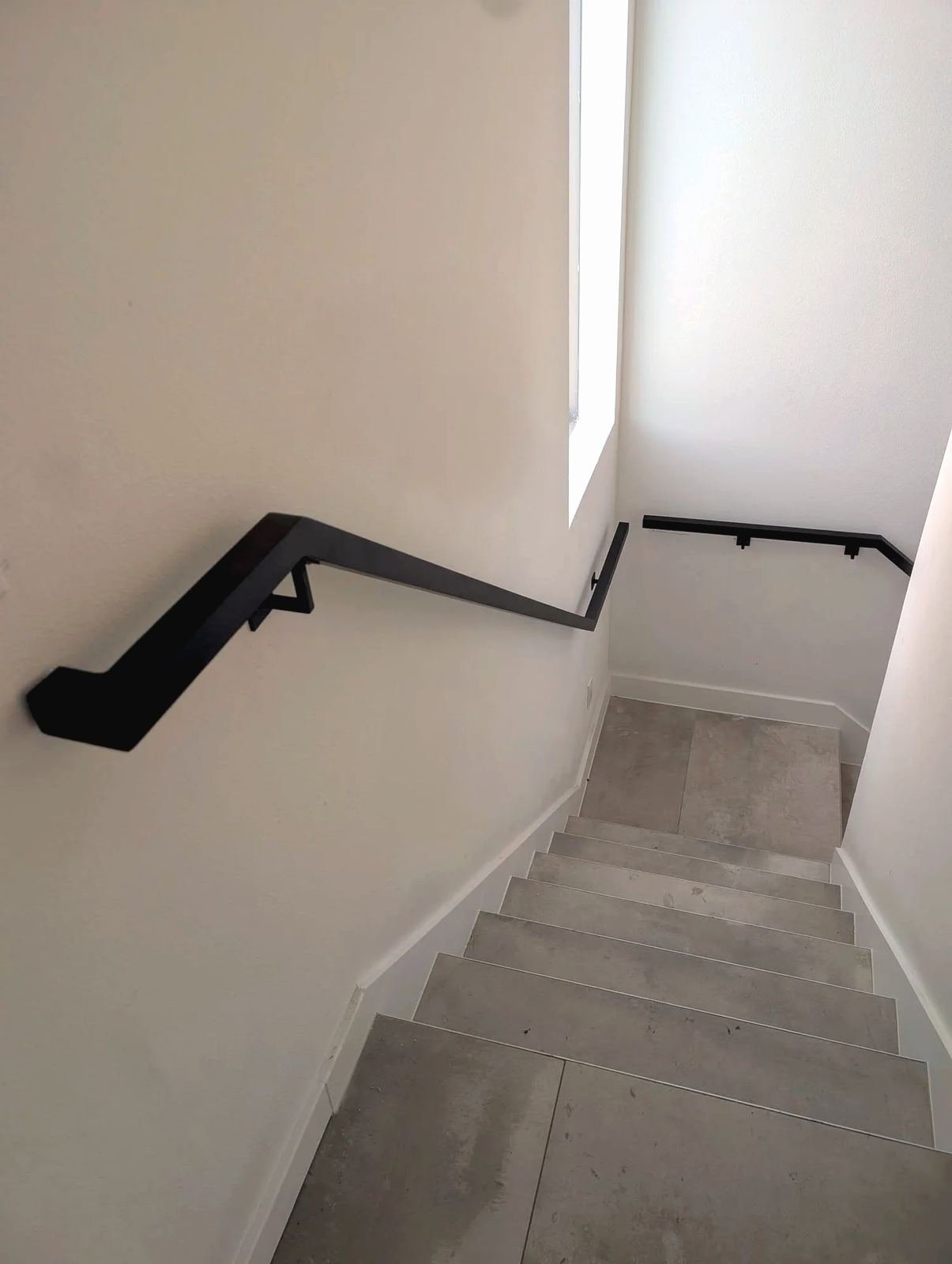 Black aluminum wall-mounted handrail turning down an interior stairway with pale tile steps and white walls