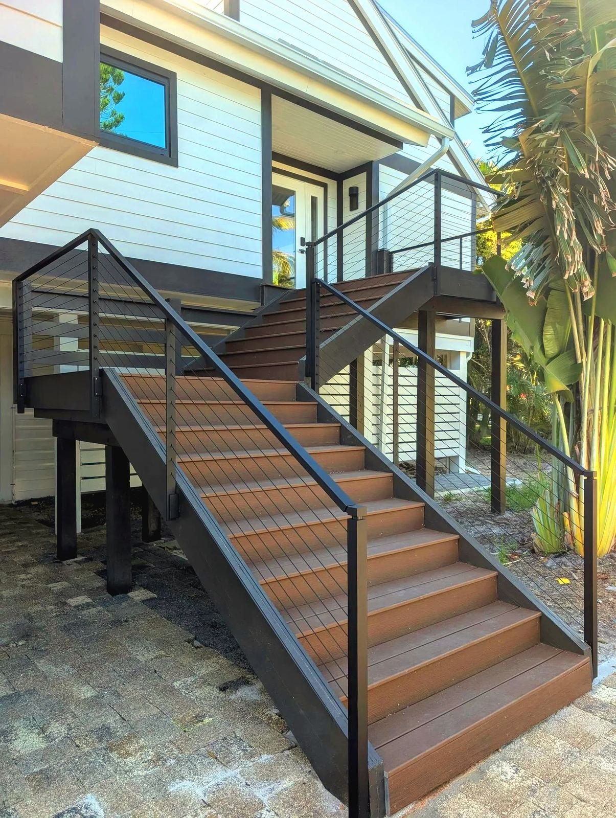 Black cable railing on a wood exterior staircase up to a white stilt house, paver landing and palms