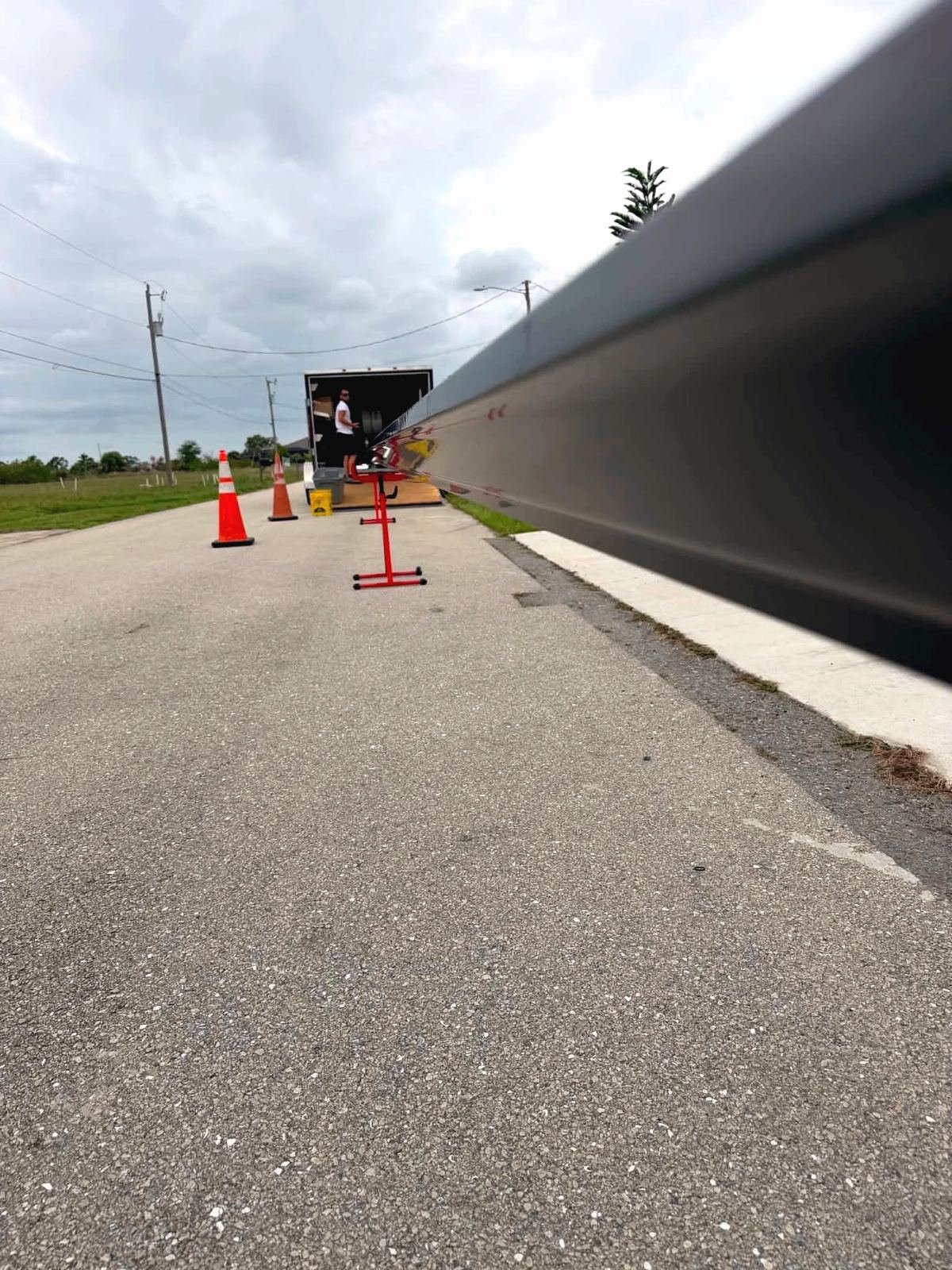 Looking down a freshly rolled dark gutter run toward the gutter machine trailer, cones and a stand on the road, overcast sky