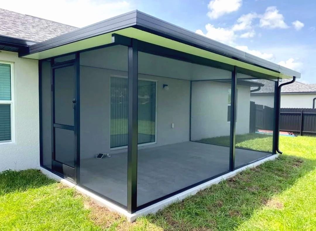 Bronze aluminum solid-roof enclosure with screen walls on a gray stucco house, bare concrete slab inside, gutter along the edge