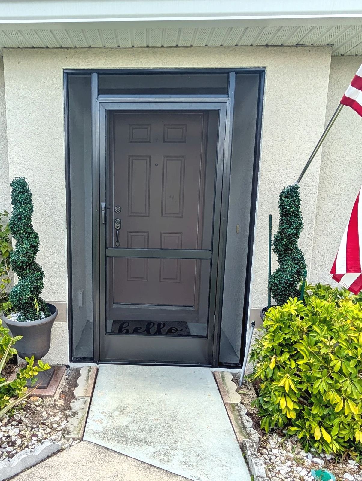 Bronze aluminum screened entry enclosure on a stucco front door, flanked by potted spiral topiaries and an American flag