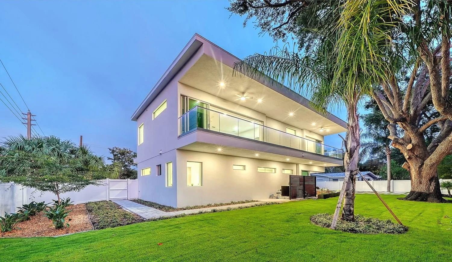 Two-story white modern house at dusk with a clean fascia line, glass balcony rail and lit soffit, palms and an oak in the yard