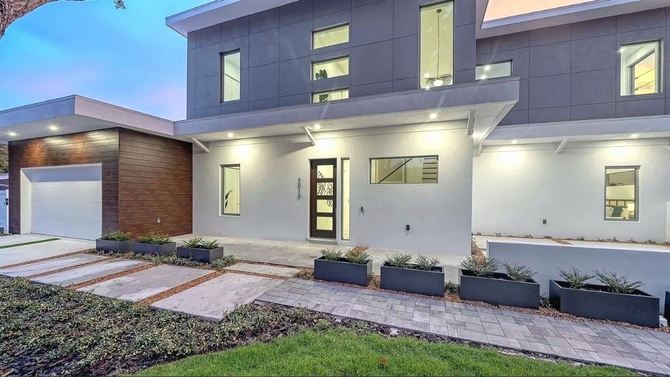 Gray two-story modern house at dusk with a wood-look cladding accent wall at the garage, lit entry and planter boxes