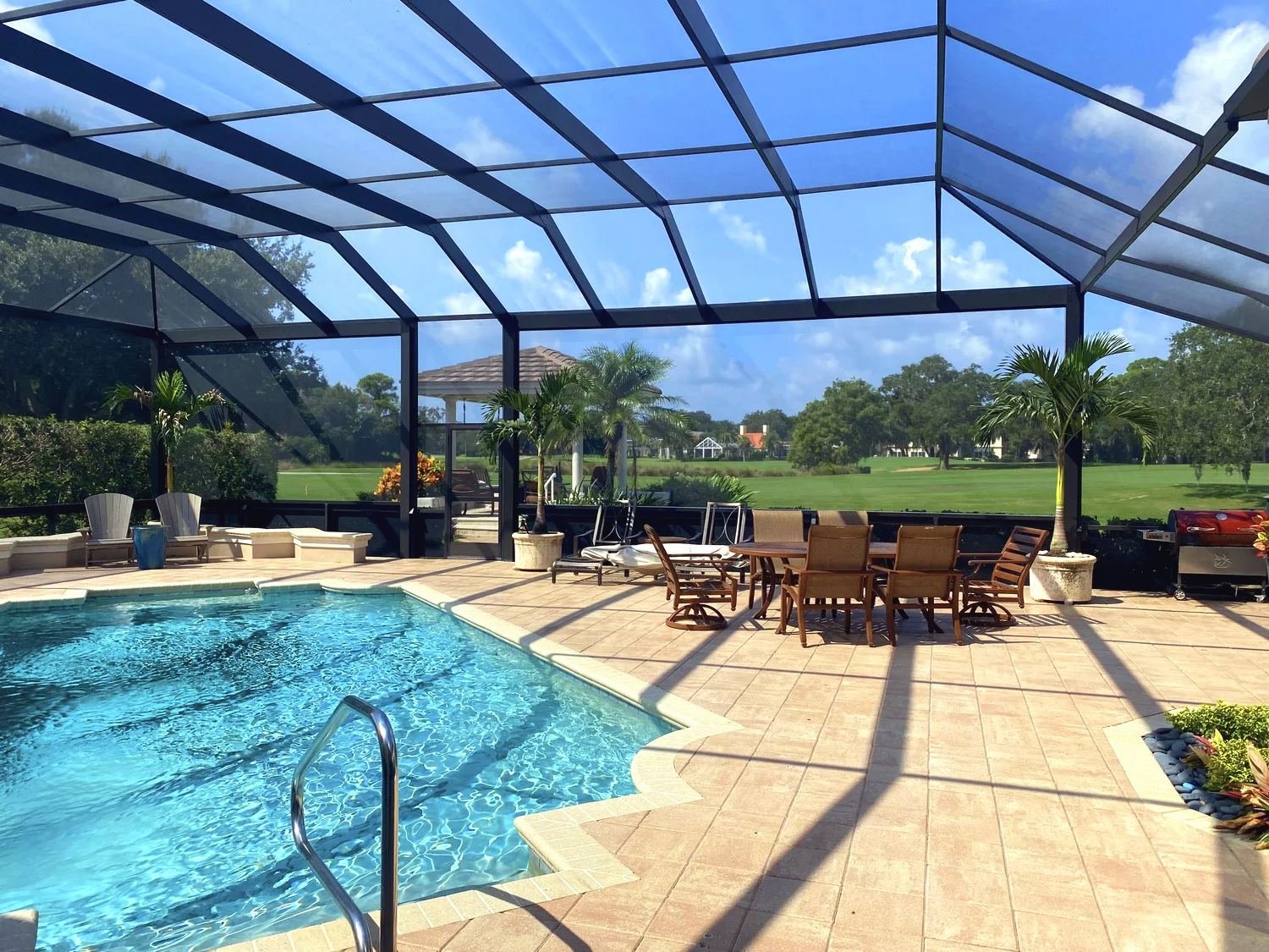 Black mansard pool cage over a pool and patio dining set, golf course fairway and palms beyond the screen