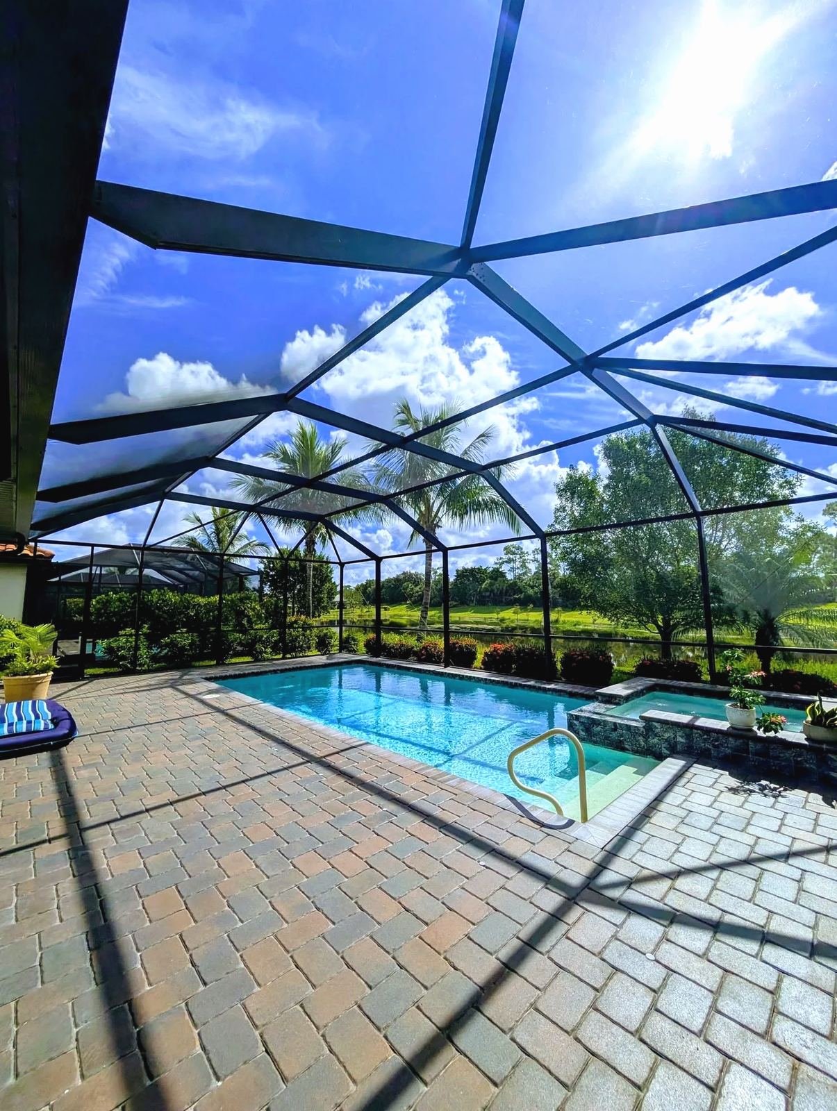 Looking up through a bronze pool cage roof at blue sky and clouds, pool and spa below, paver deck