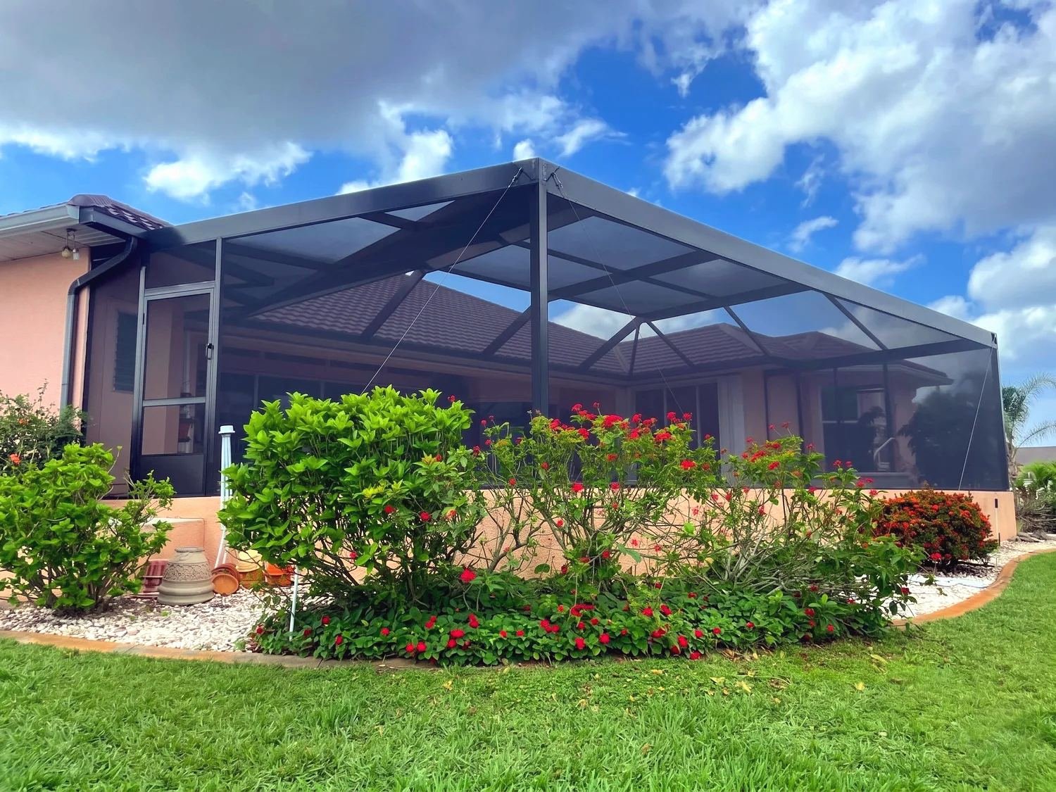 Black aluminum pool cage on the back of a pink stucco ranch house with a tile roof, red flowering shrubs and white rock beds in front, blue sky with clouds