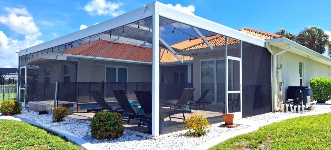 White aluminum pool cage with string lights over a small pool and lounge chairs, terracotta tile roof behind, white rock and shrubs around the base