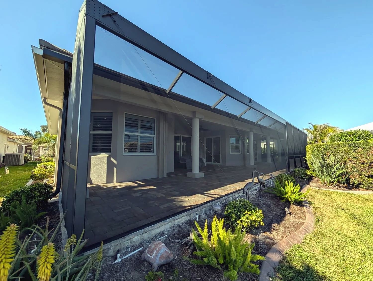 Dark bronze solid-roof patio enclosure with screen walls extending off a stucco house, paver floor and landscaping, Southwest Florida