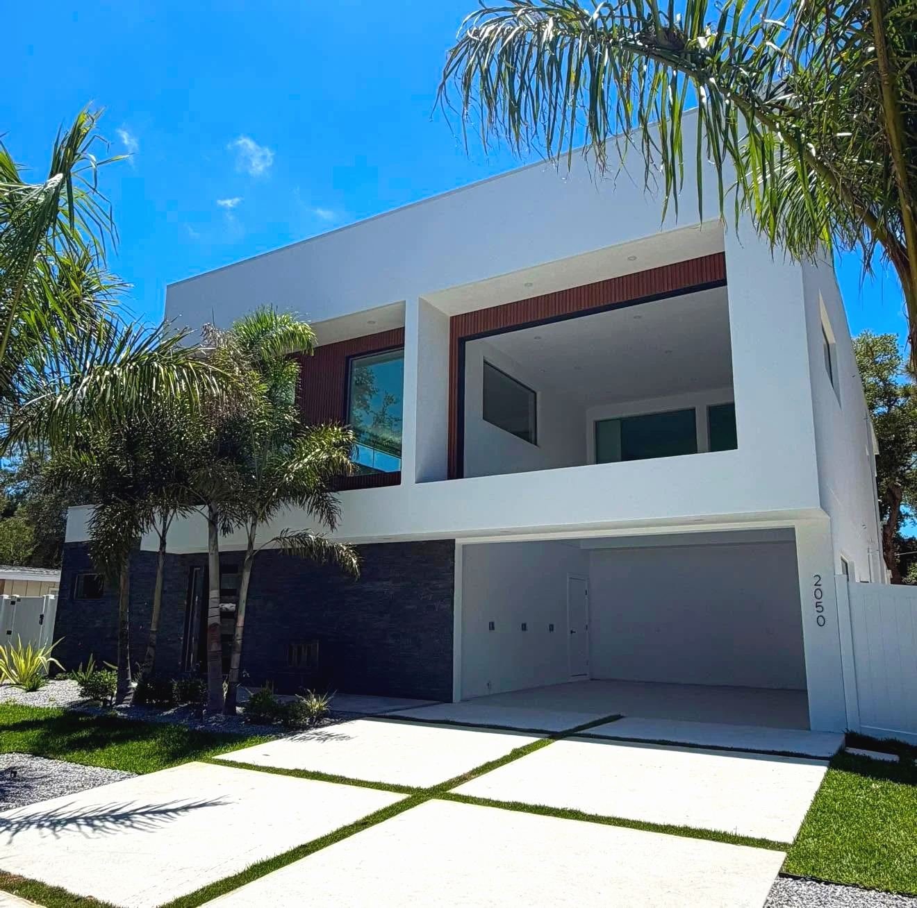 Two-story white modern house with wood-look aluminum cladding around the second-floor windows, dark stone at the garage, palms and a paver driveway