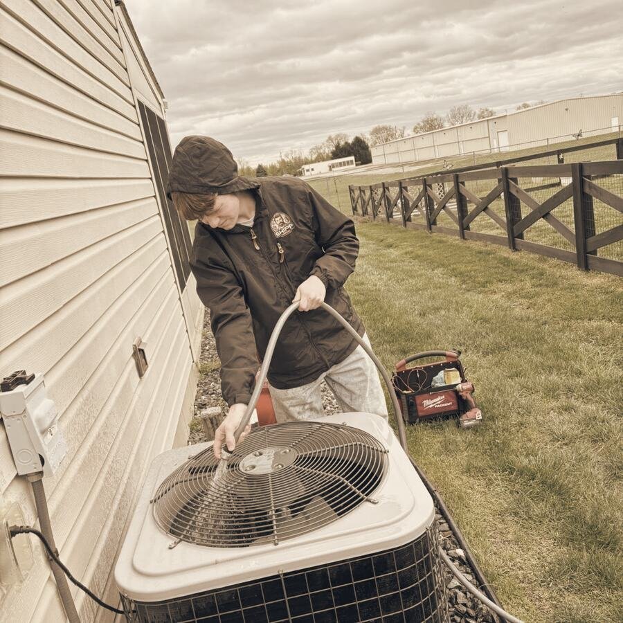 Shipp's tech in a SHIPP hoodie cleaning an outdoor AC unit beside a black board fence